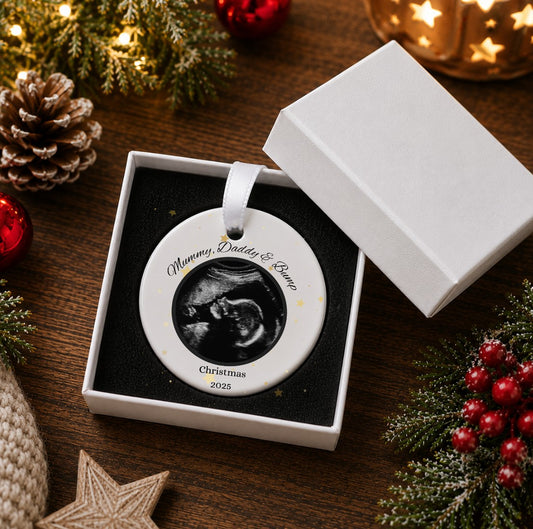 A personalised round ceramic Christmas ornament displayed in an open white gift box on a dark wooden table, surrounded by festive holiday decorations including pine branches, red berries, a pinecone, and warm string lights. The ornament features a black-and-white ultrasound photo in the centre with the custom text 'Mummy, Daddy & Bump' and 'Christmas 2026' printed on a white background.