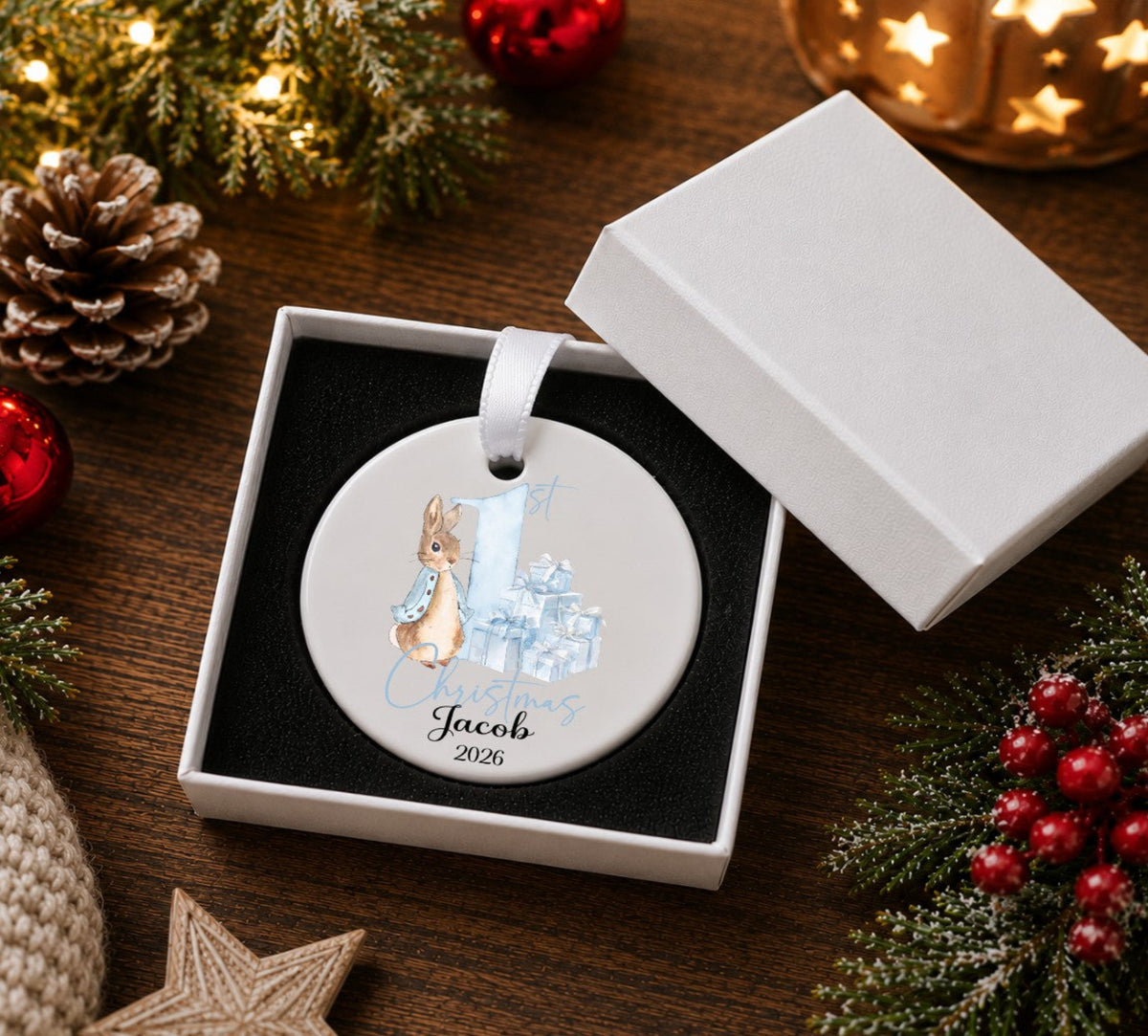 A personalised round Christmas ornament displayed in an open white gift box. The ornament features an illustration of a rabbit and gift box with the name 'Jacob' and year '2026' printed below.