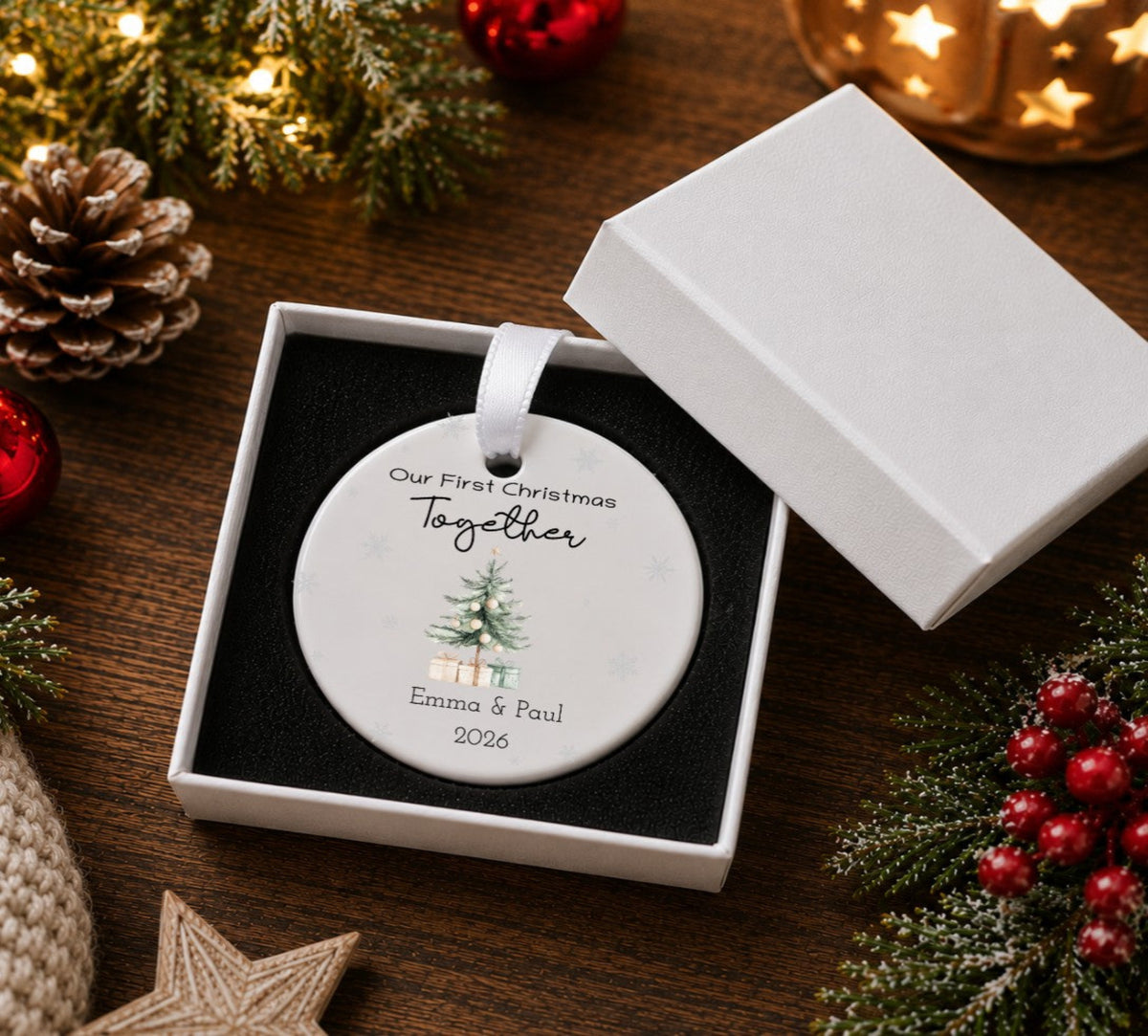 A personalised round Christmas ornament displayed in an open white gift box on a dark wooden surface, surrounded by festive decorations including pine branches, red berries, a pinecone, and a glowing star-lit lantern. The ornament is printed with the text 'Our First Christmas Together', a small Christmas tree illustration, and the names 'Emma & Paul' with the year '2026'.