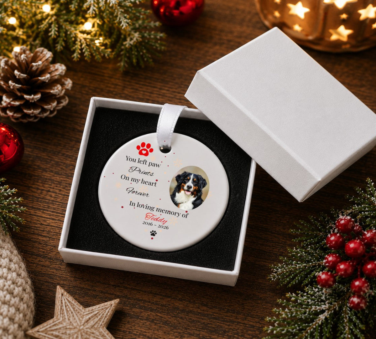 A personalised round white pet memorial Christmas ornament in a white gift box The ornament features a photo of a Bernese mountain dog, red paw print graphics, and printed text reading 'You left paw Prints On my heart Forever In loving memory of Teddy 2016 - 2026'.