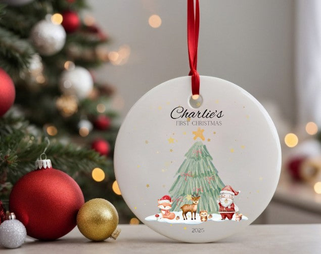 Personalised Christmas ornament with 'Charlie's First Christmas' text and festive woodland scene , hanging in front of a decorated tree.