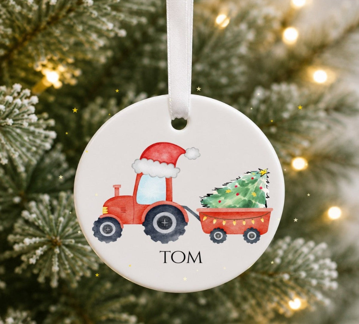 A round white ceramic Christmas ornament hanging from a white ribbon, featuring a red tractor with a Santa hat pulling a trailer with a Christmas tree.