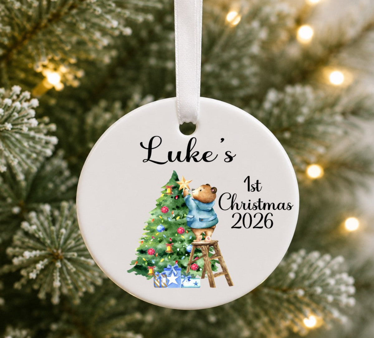 A round white ceramic Christmas ornament hanging on a tree, printed with the name 'Luke's', '1st Christmas 2026', and an illustration of a teddy bear decorating a Christmas tree.