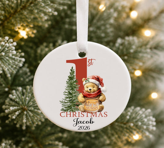 A round white ceramic Christmas ornament hanging from a white ribbon, featuring a teddy bear in a Santa hat next to a small tree and the text '1st Christmas Jacob 2026'. The ornament is photographed against a blurred Christmas tree with warm string lights.