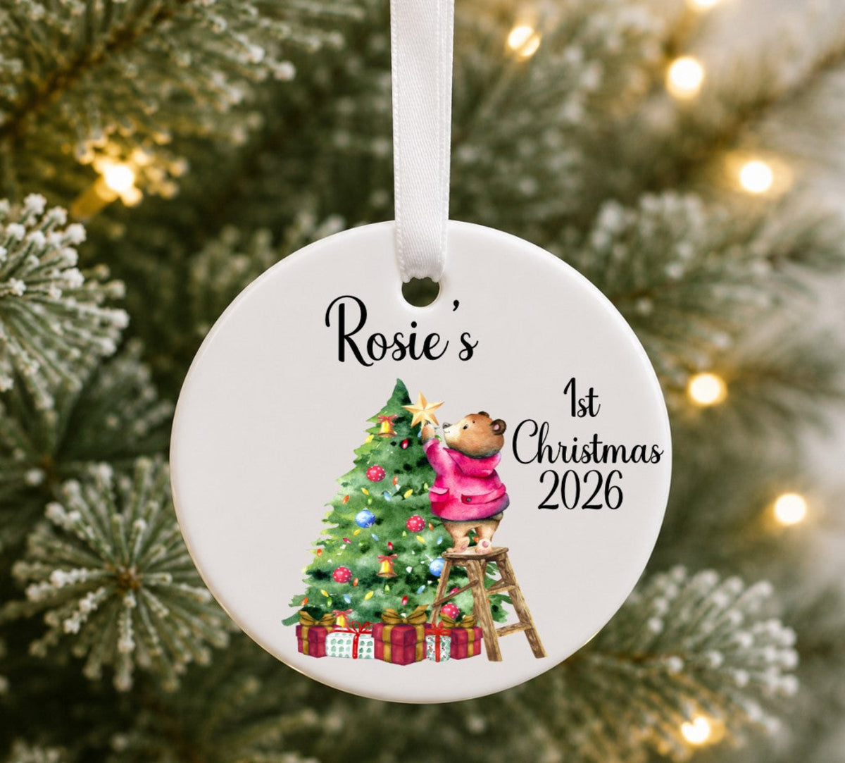 A round white ceramic Christmas ornament hanging on a tree, printed with the name 'Rosie's', '1st Christmas 2026', and an illustration of a teddy bear decorating a Christmas tree.