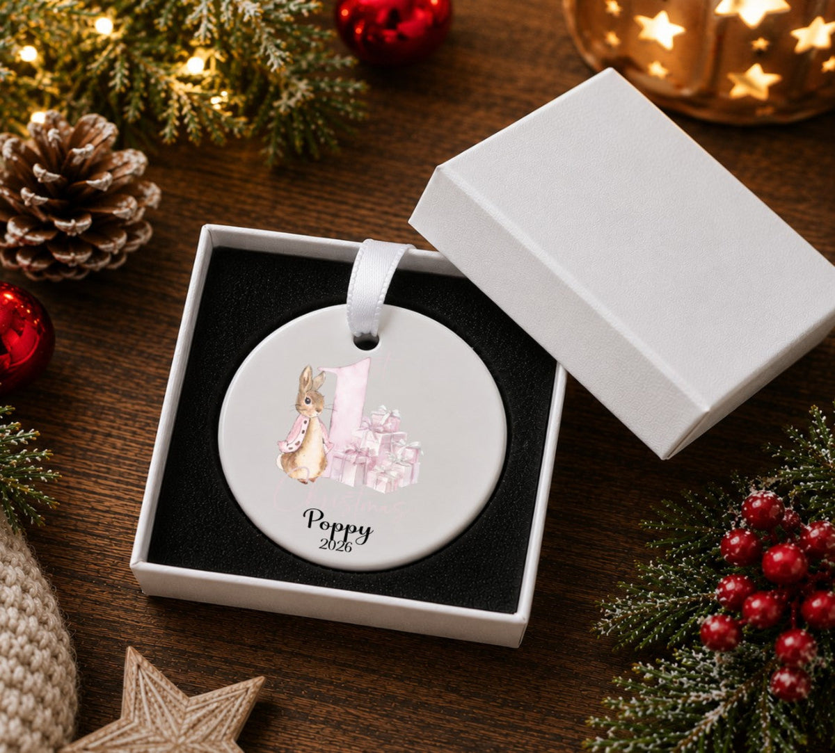 A round white personalised Christmas ornament with a printed illustration of a rabbit and gift boxes, displayed inside an open white gift box on a dark wooden table surrounded by holiday decorations.