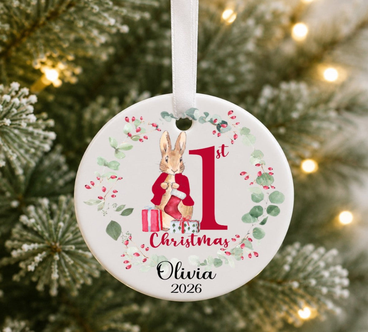 Ceramic round Christmas ornament hanging on a tree with an illustration of a rabbit wearing a red jacket, personalised with 1St Christmas Olivia 2026.