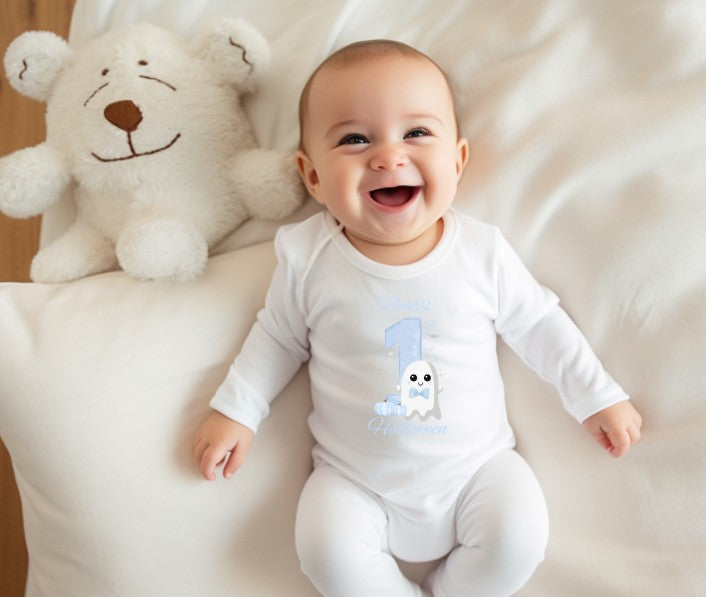 Baby wearing a white onesie with a blue number '1' and ghost design, sitting on a white surface with a teddy bear.