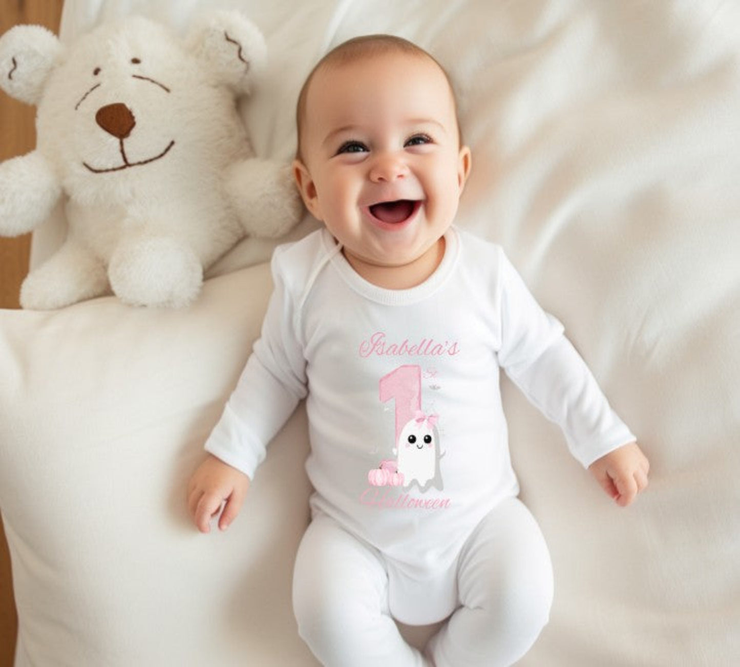 Baby wearing a white onesie with pink text and graphics, sitting on a soft surface with a teddy bear.