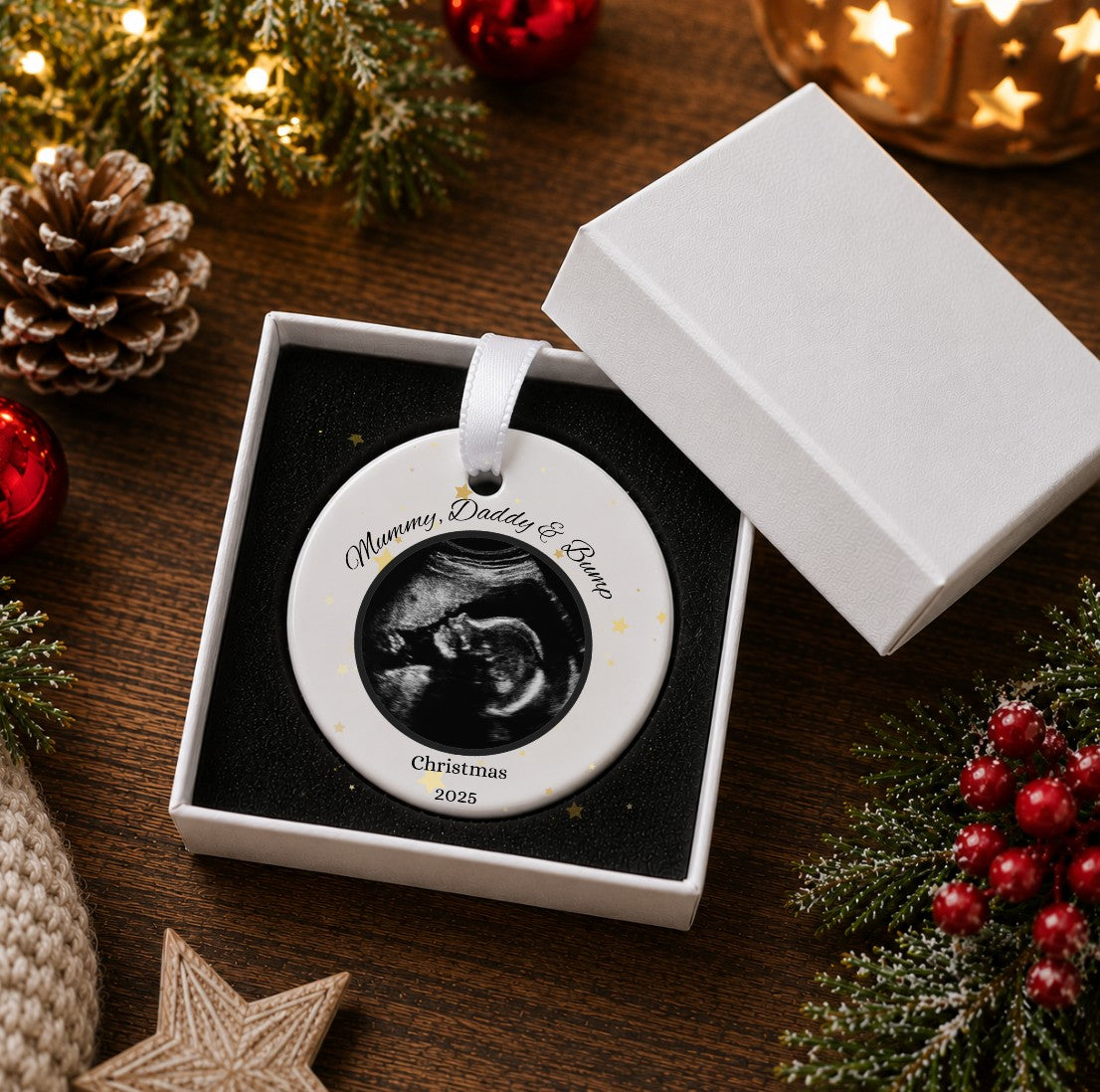 A personalised round ceramic Christmas ornament displayed in an open white gift box on a dark wooden table, surrounded by festive holiday decorations including pine branches, red berries, a pinecone, and warm string lights. The ornament features a black-and-white ultrasound photo in the centre with the custom text 'Mummy, Daddy & Bump' and 'Christmas 2026' printed on a white background.