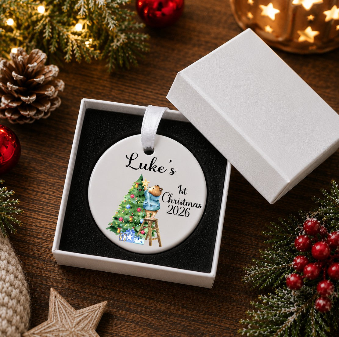 A round white ceramic Christmas ornament presented in a gift box, printed with the name 'Luke's', '1st Christmas 2026', and an illustration of a teddy bear decorating a Christmas tree.