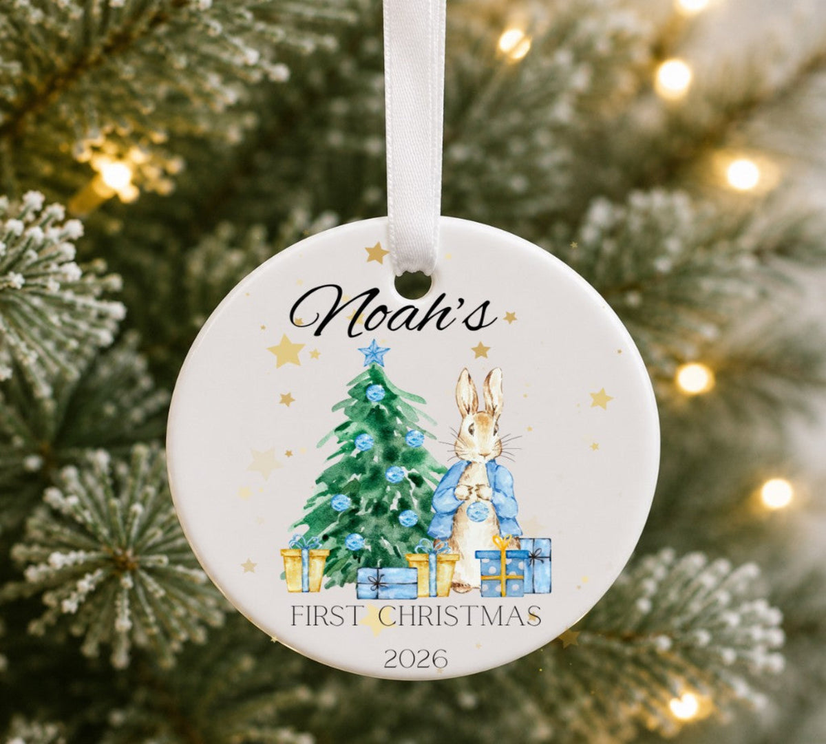 A personalised round ceramic Christmas ornament hanging from a white ribbon, featuring an illustration of Peter Rabbit and a decorated Christmas tree with the text 'Noah's FIRST CHRISTMAS 2026'. The ornament is photographed in front of a blurred Christmas tree with warm string lights.