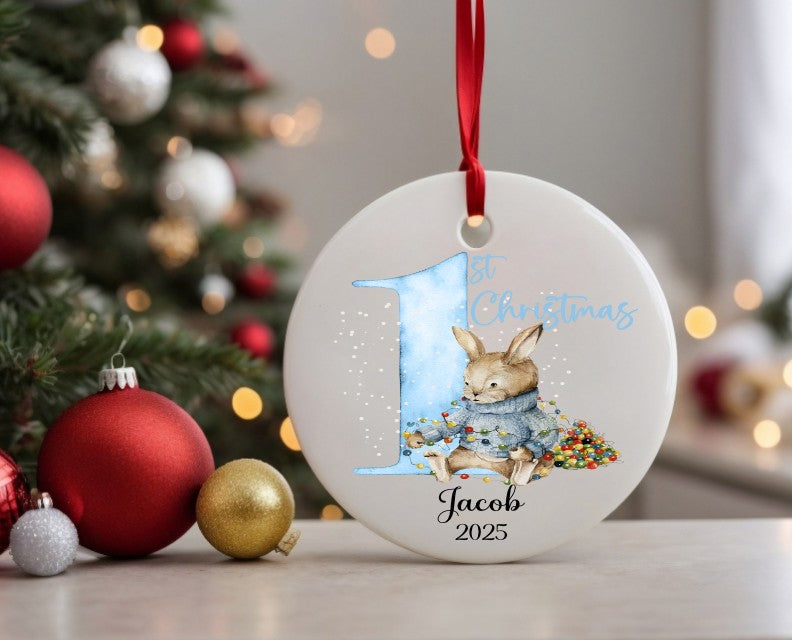 Personalised Christmas ornament with a rabbit and '1st Christmas' text, decorated tree in the background.