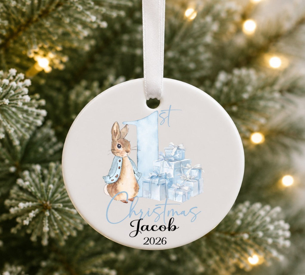 A personalised round ceramic Christmas ornament hanging from a white ribbon, featuring a watercolour illustration of Peter Rabbit, a large blue number 1, wrapped presents, and the text '1st Christmas Jacob 2026', set against a blurred Christmas tree background with warm lights.