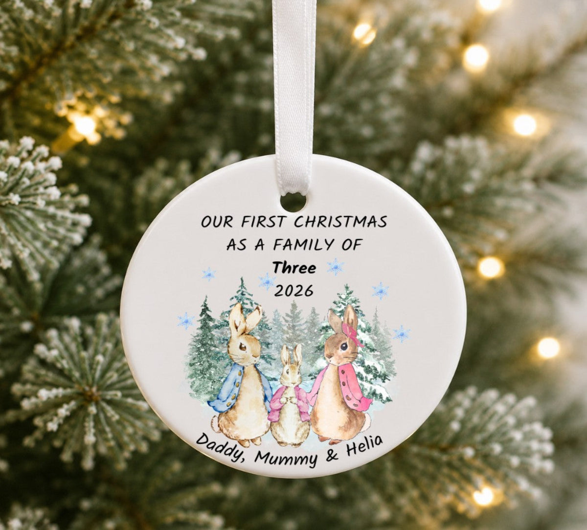 A round white ceramic Christmas ornament hanging on a Christmas tree with warm string lights. The ornament is printed with a family of rabbits illustration and the text 'OUR FIRST CHRISTMAS AS A FAMILY OF Three 2026' and 'Daddy, Mummy & Helio'.