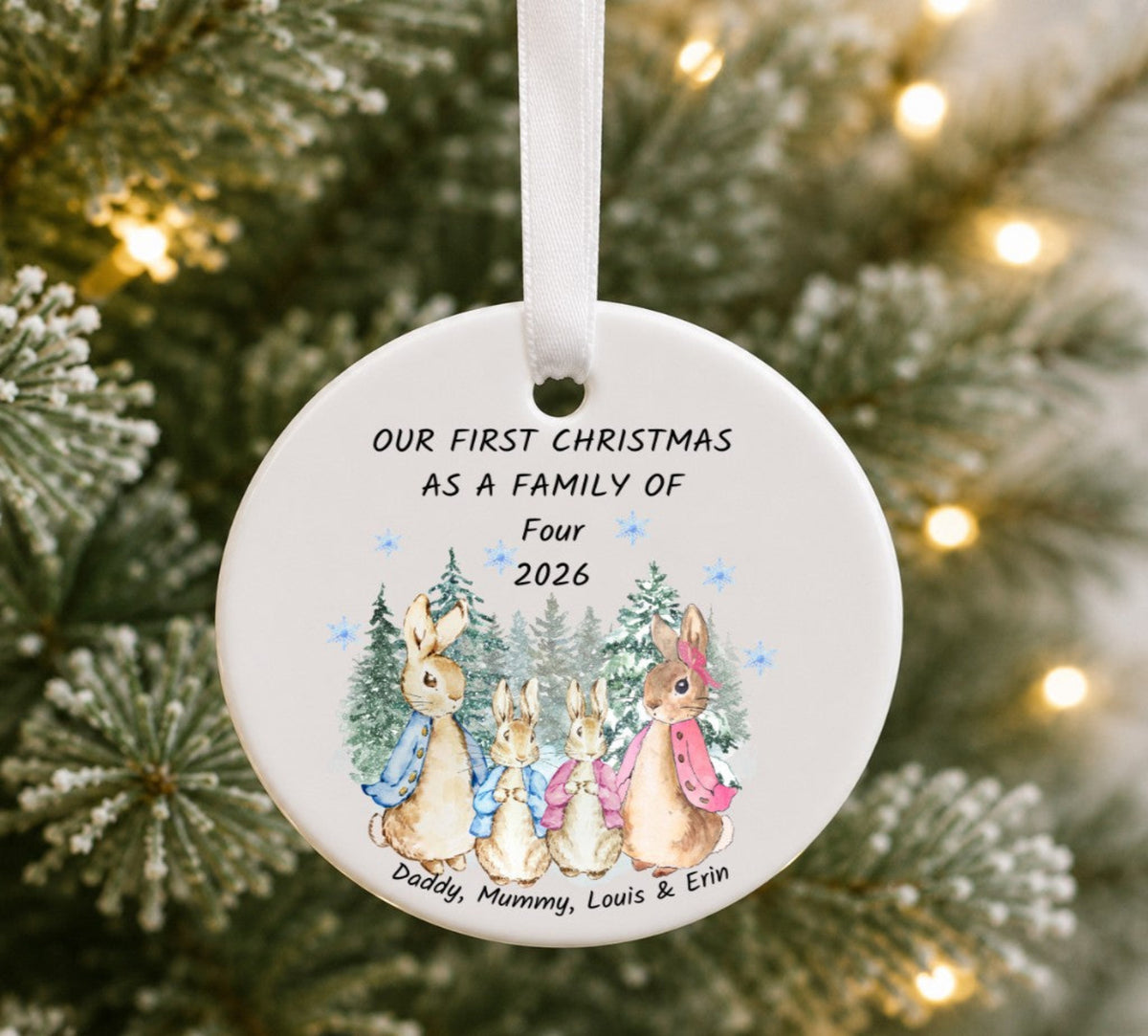 A personalized round ceramic Christmas ornament hanging on a blurred Christmas tree with warm lights. The white ornament features a printed illustration of three Peter Rabbit characters and the text 'OUR FIRST CHRISTMAS AS A FAMILY of Four 2026' with names at the bottom.