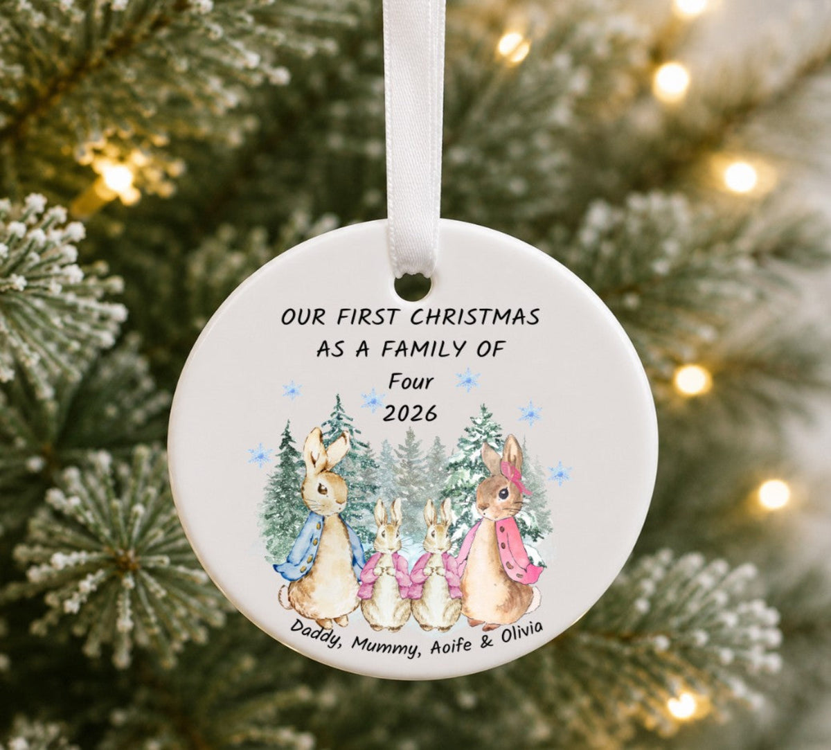 A round white ceramic Christmas ornament hanging from a white ribbon, printed with the text 'OUR FIRST CHRISTMAS AS A FAMILY OF Four 2026' and an illustration of three rabbits among pine trees. The ornament is photographed in front of a blurred Christmas tree with warm string lights.