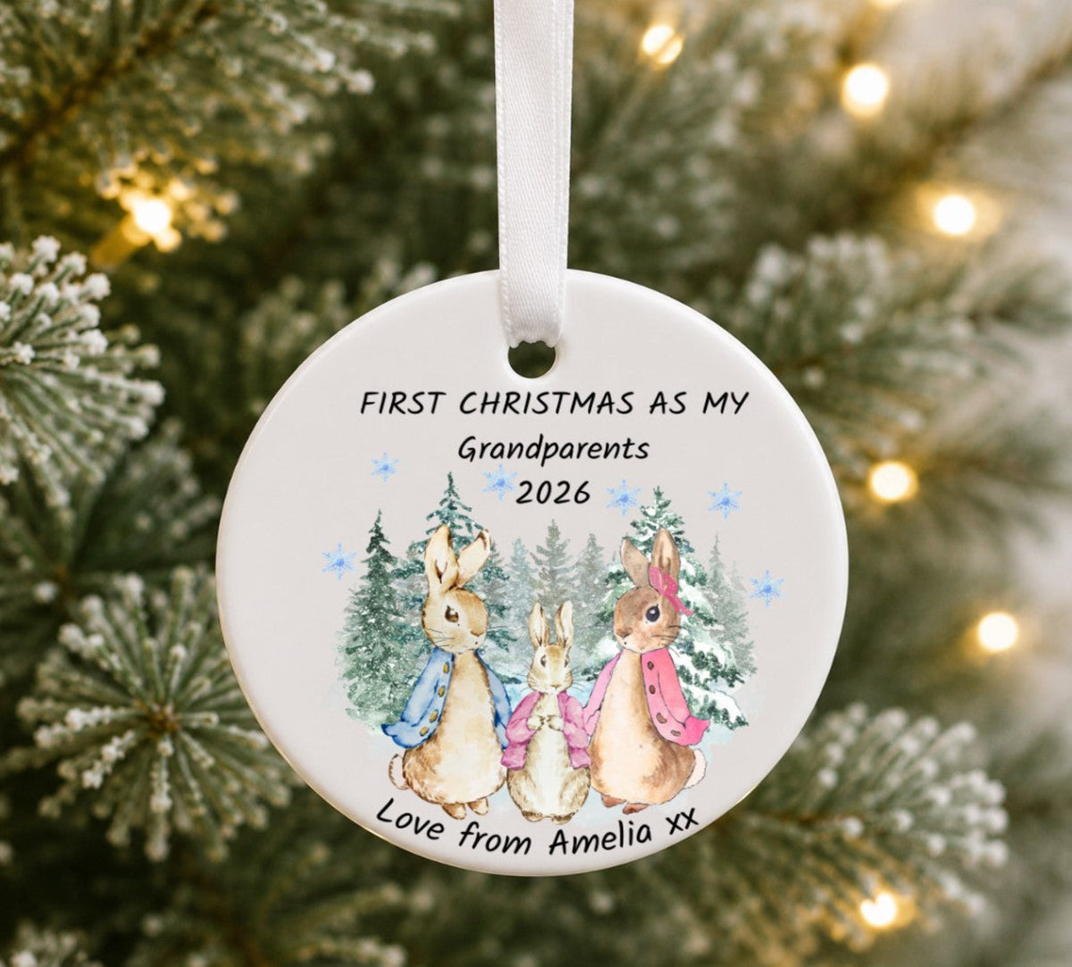 A round white ceramic Christmas ornament hanging from a white ribbon, featuring a painted illustration of three rabbits in front of pine trees, with the text 'FIRST CHRISTMAS AS MY Grandparents 2026' and 'Love from Amelia xx'. The ornament is photographed in front of a blurred Christmas tree with warm lights.