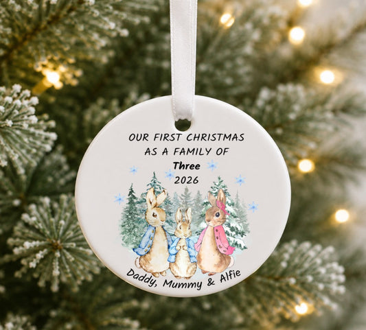 A personalised round ceramic Christmas ornament hanging on a Christmas tree. The white ornament features a printed illustration of three rabbits in blue and pink jackets under snowy trees, with the text 'OUR FIRST CHRISTMAS AS A FAMILY OF Three 2026' and 'Daddy, Mummy & Alfie'.