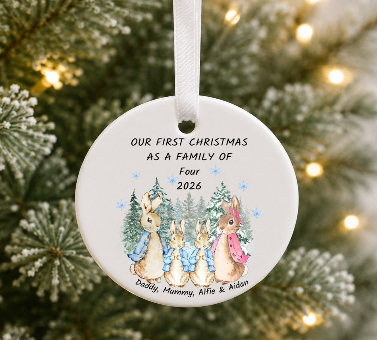 A round white ceramic Christmas ornament hanging from a white ribbon, printed with the text 'OUR FIRST CHRISTMAS AS A FAMILY OF Four 2026' and an illustration of four rabbits among pine trees. The ornament is photographed hanging on a Christmas tree with blurred lights in the background.