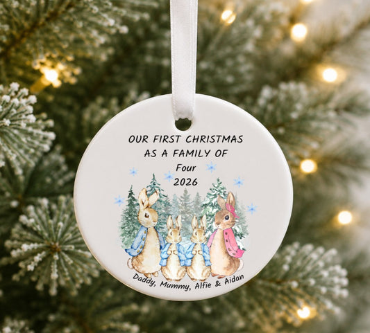A round white ceramic Christmas ornament hanging from a white ribbon, printed with the text 'OUR FIRST CHRISTMAS AS A FAMILY OF Four 2026' and an illustration of four rabbits among pine trees. The ornament is photographed hanging on a Christmas tree with blurred lights in the background.