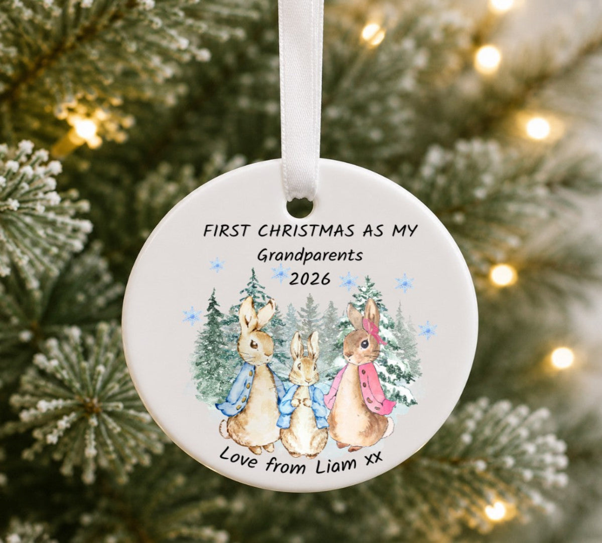 A round white ceramic Christmas ornament hanging from a white ribbon, featuring a printed illustration of three rabbits in winter clothing with the text 'FIRST CHRISTMAS AS MY Grandparents 2026' and 'Love from Liam xx'. The ornament is photographed against a blurred Christmas tree background with warm string lights.