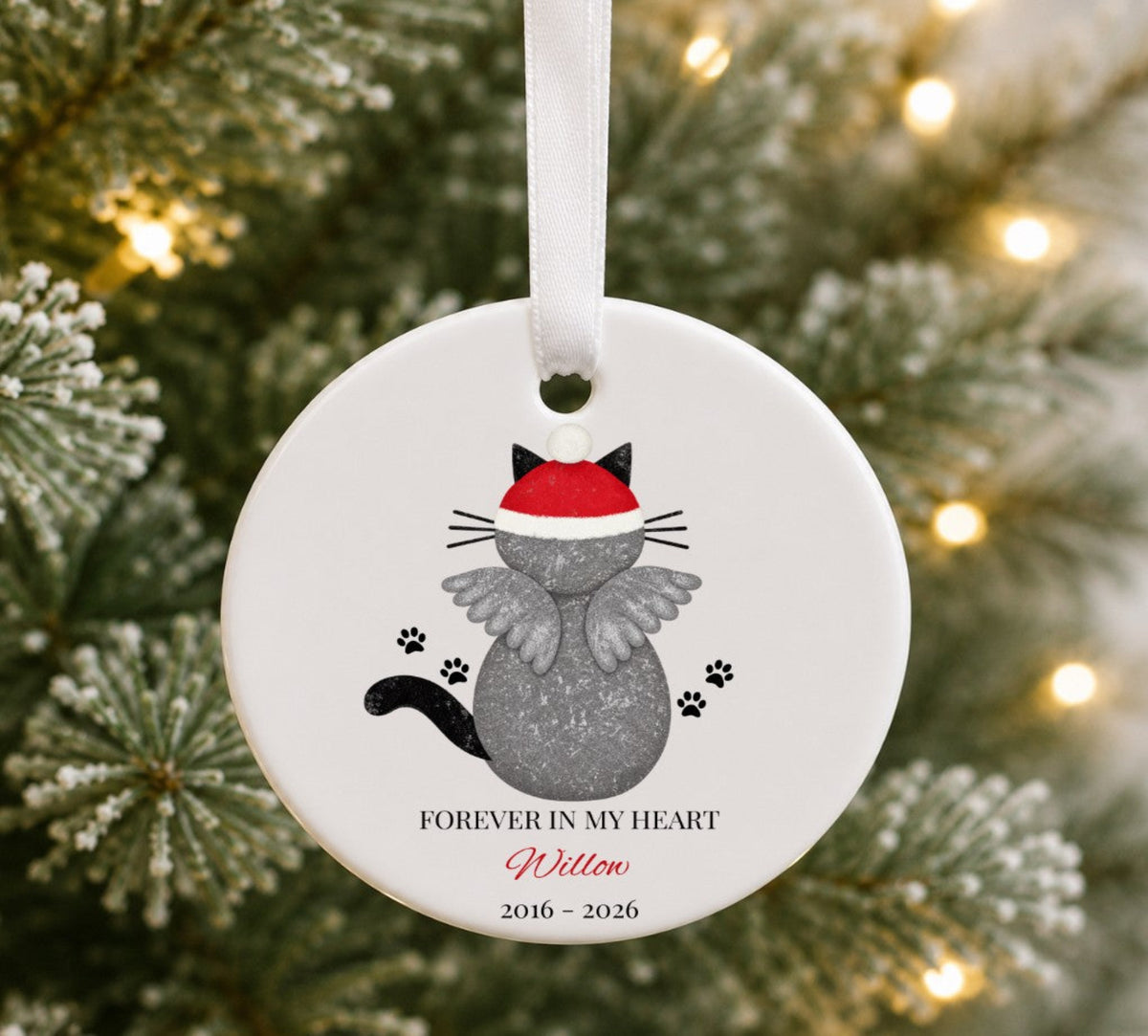 A round white ceramic Christmas ornament hanging from a white ribbon, featuring a grey cat with angel wings and a red Santa hat, with the text 'FOREVER IN MY HEART Willow 2016 - 2026' on a blurred Christmas tree background.