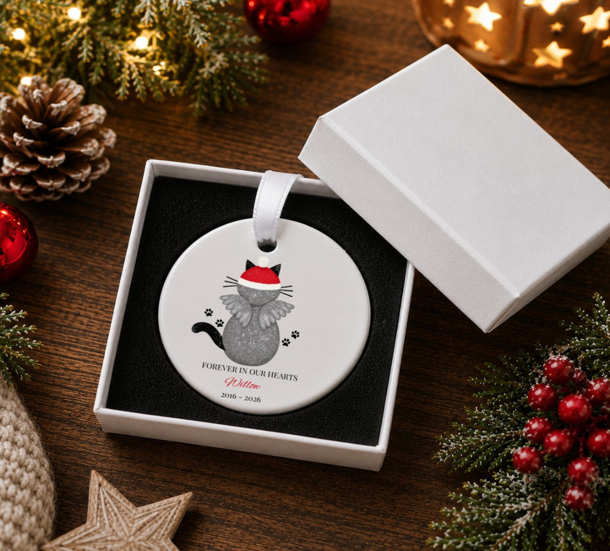 A round white Christmas ornament with a gray cat wearing a red Santa hat, displayed in an open white gift box on a dark wooden table surrounded by holiday decorations.