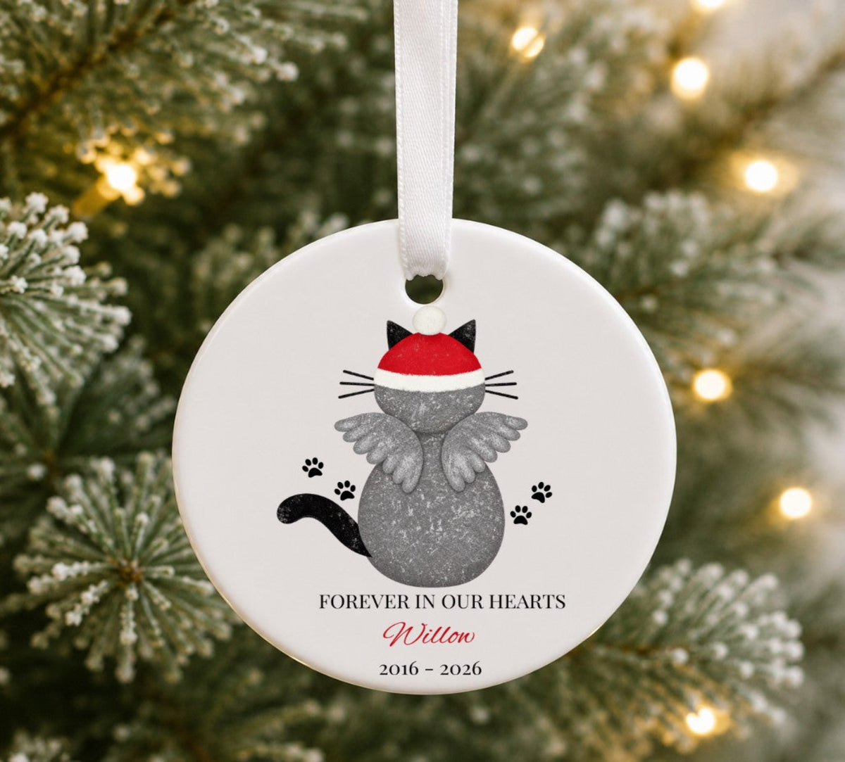 A round white ceramic Christmas ornament hanging from a white ribbon, featuring a grey cat wearing a red Santa hat, with the text 'FOREVER IN OUR HEARTS Willow 2016 - 2026' on a blurred Christmas tree background with warm lights.