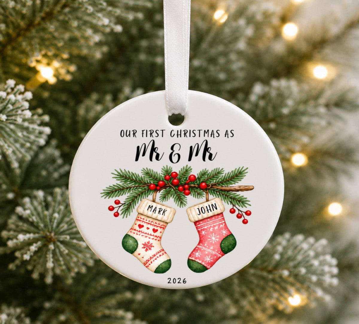 A round white ceramic Christmas ornament hanging from a white ribbon, printed with the text 'OUR FIRST CHRISTMAS AS Mr & Mr' above an illustration of two personalized Christmas stockings labelled 'MARK' and 'JOHN' with green pine branches and red berries, set against a blurred Christmas tree background with warm lights.