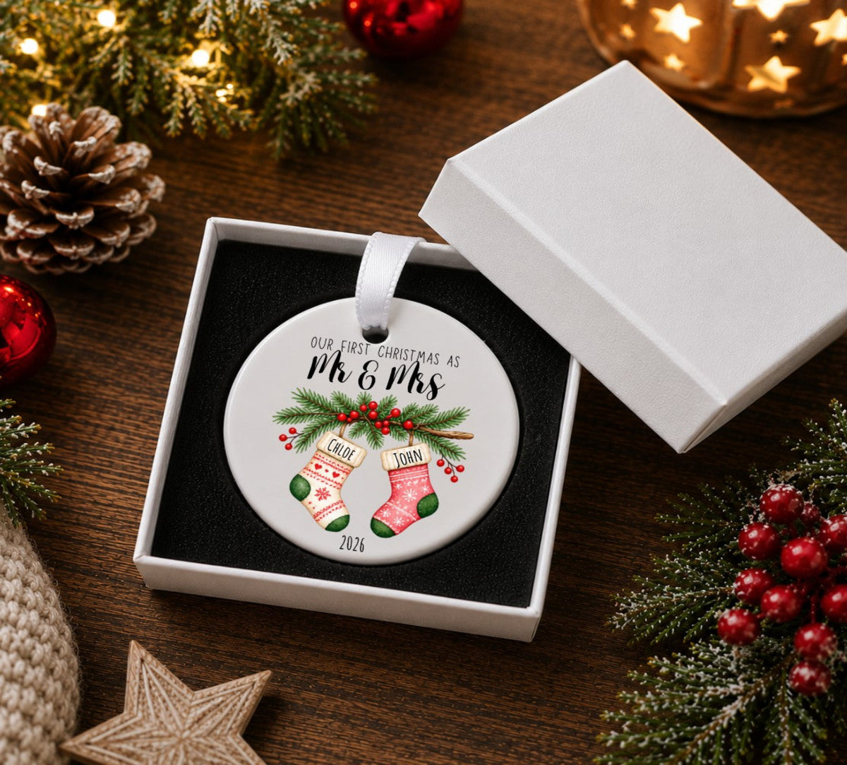 A personalised round Christmas ornament displayed in an open white gift box on a wooden table, surrounded by holiday decorations including pine branches, red berries, a pinecone, and warm string lights. The ornament features a white background with black script text reading 'OUR FIRST CHRISTMAS AS MR & MRS' above an illustration of two stockings with green holly and red berries.