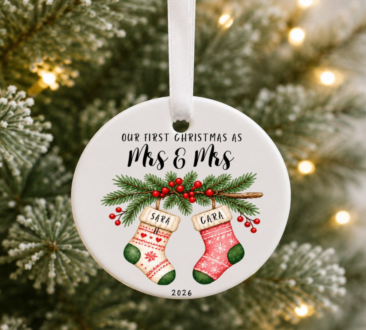 A round white ceramic Christmas ornament hanging from a white ribbon, printed with the text 'OUR FIRST CHRISTMAS AS Mrs & Mrs ' above two illustrated stockings, green pine branches, and red berries, with blurred evergreen branches and warm string lights in the background.