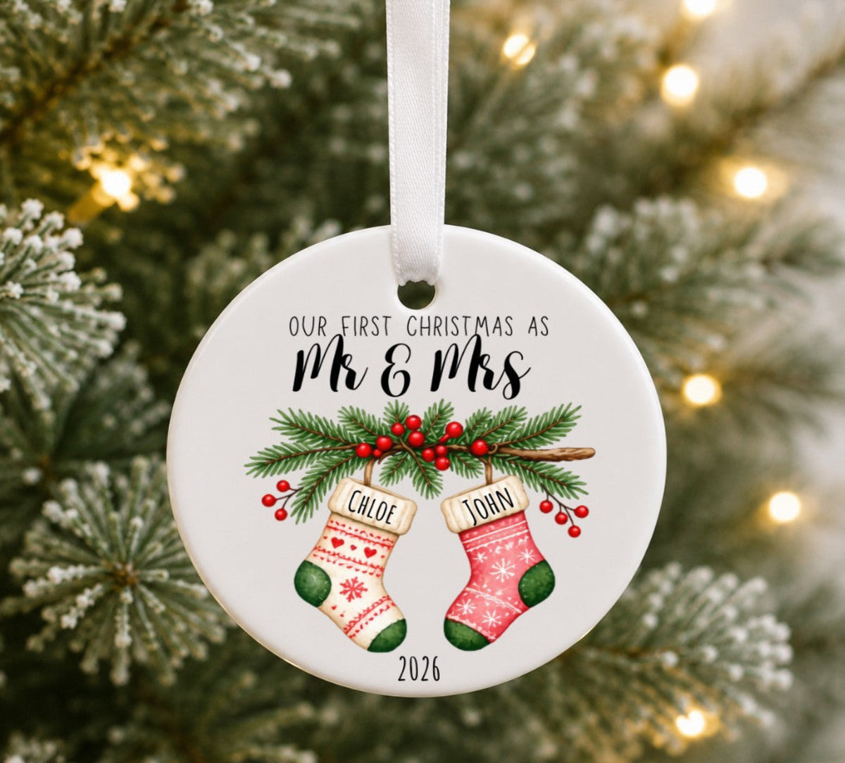 A round white ceramic Christmas ornament hanging from a white ribbon, printed with the text 'OUR FIRST CHRISTMAS AS Mr & Mrs' above an illustration of two personalized stockings named Chloe and John with green pine branches and red berries, set against a blurred Christmas tree background.