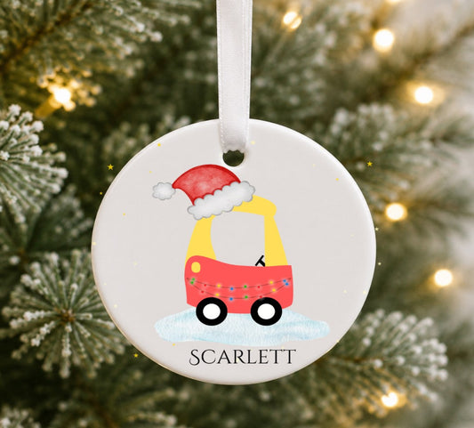 A personalised round ceramic Christmas ornament hanging on a blurred Christmas tree with warm lights. The ornament is white and features a cartoon toy car in yellow, red, and blue with a red Santa hat, the name 'SCARLETT' printed at the bottom, and a white hanging ribbon.
