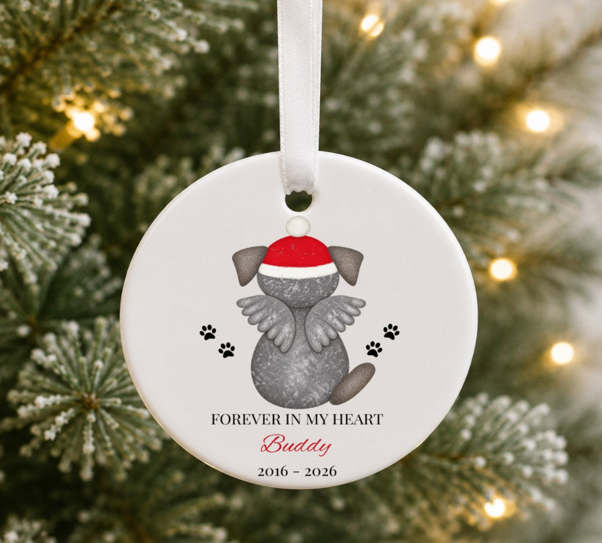 A round white pet memorial Christmas ornament hanging from a white ribbon, featuring a grey angel dog wearing a red Santa hat with the text 'FOREVER IN MY HEART Buddy 2016 - 2026' against a blurred Christmas tree background.
