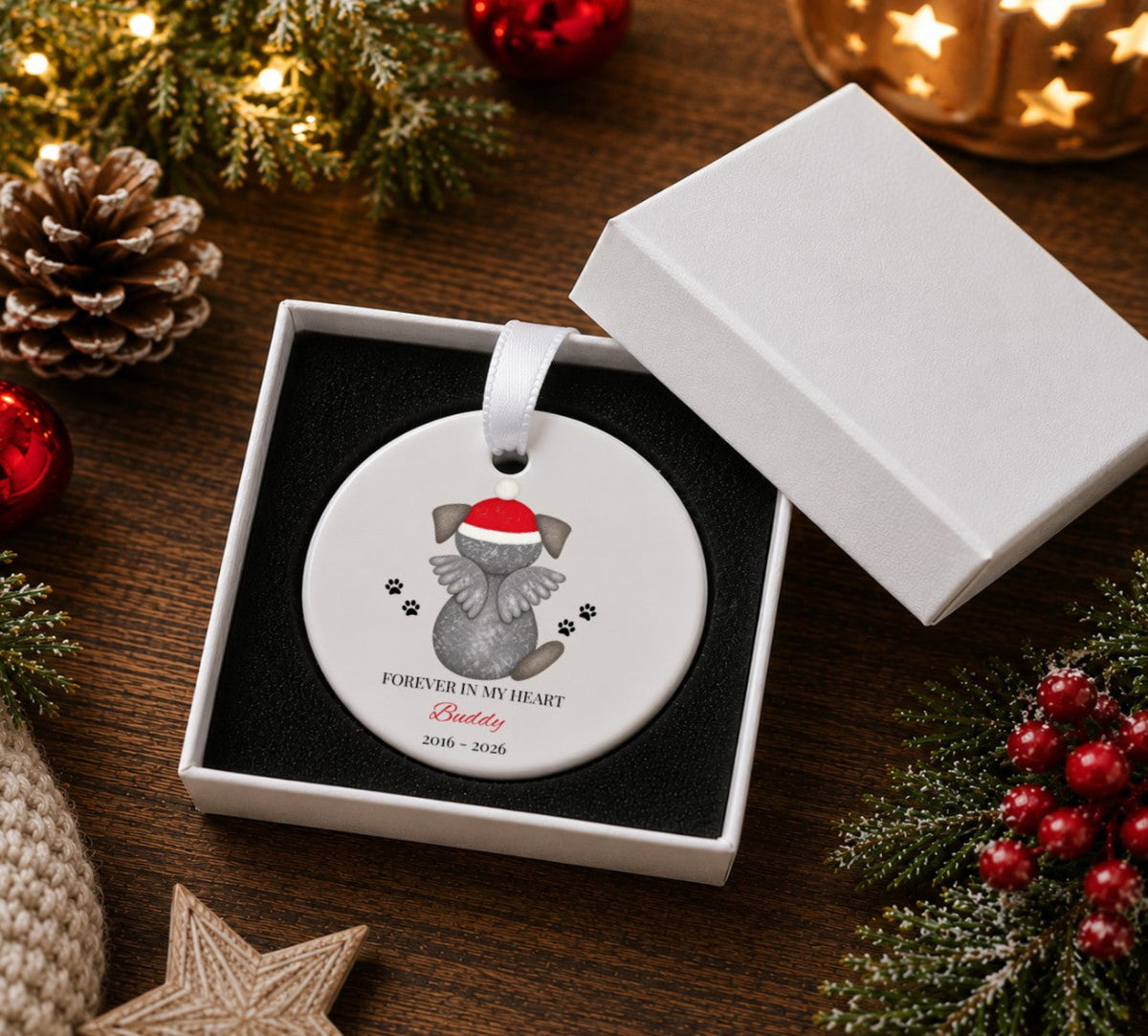 A round white Christmas ornament in a white gift box, featuring a grey dog wearing a red Santa hat with the text 'FOREVER IN MY HEART Buddy 2016 - 2026'. The box is open on a wooden table surrounded by holiday decorations like pinecones, evergreen branches, and red berries.