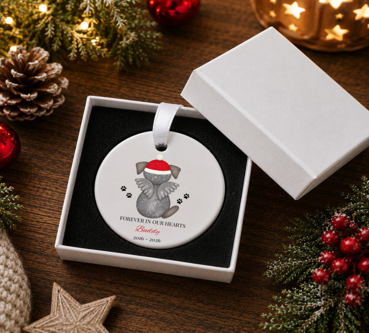 A round white pet memorial Christmas ornament displayed in an open white gift box on a wooden table, surrounded by holiday decorations including pine branches, red berries, a pinecone, and a glowing star candle. The ornament features a grey dog wearing a red Santa hat with black paw prints and the text 'FOREVER IN OUR HEARTS Betty 2018 - 2026'.