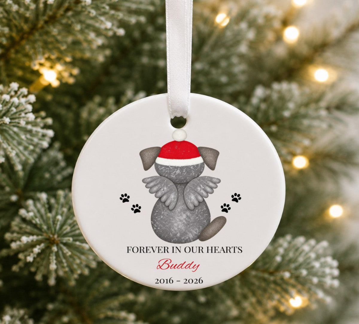 A round white pet memorial Christmas ornament hanging from a white ribbon, featuring a grey dog illustration with angel wings and a red Santa hat, with the text 'FOREVER IN OUR HEARTS Buddy 2016 - 2026' against a blurred Christmas tree background.