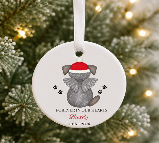 A round white pet memorial Christmas ornament hanging from a white ribbon, featuring a grey dog illustration with angel wings and a red Santa hat, with the text 'FOREVER IN OUR HEARTS Buddy 2016 - 2026' against a blurred Christmas tree background.