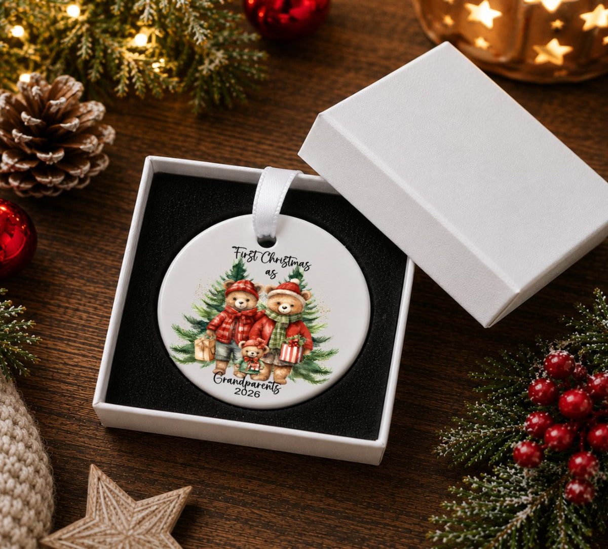 A round white Christmas ornament with a printed illustration of two teddy bears in red holiday clothes, displayed inside an open white gift box on a dark wooden table surrounded by festive decorations like pinecones, evergreen branches, red berries, and warm string lights.