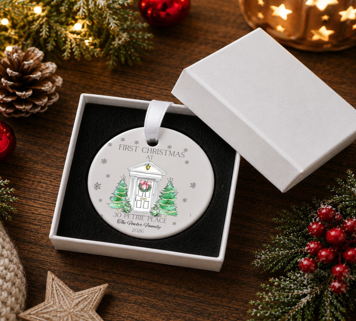 A round white ceramic Christmas ornament displayed in an open white gift box . The ornament features a printed illustration of a white house with a red wreath and green trees, with the text 'FIRST CHRISTMAS' and '30 PETITE PLACE 2026.