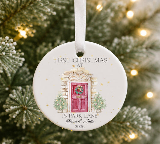 A personalised round ceramic Christmas ornament hanging from a white ribbon, featuring an illustration of a red front door with a wreath and the text 'FIRST CHRISTMAS AT 15 PARK LANE Paul & Julie 2026'. 