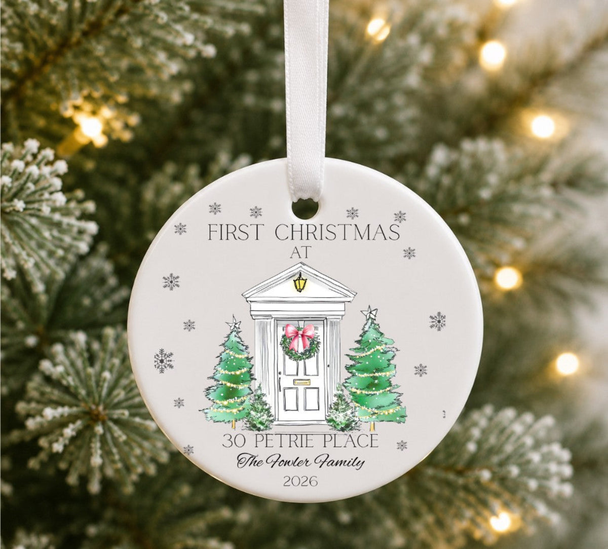 A round white ceramic Christmas ornament hanging in front of a blurred Christmas tree with warm lights. The ornament is printed with a festive illustration of a house door, wreath, two green Christmas trees, and the text 'FIRST CHRISTMAS AT 30 PETRIE PLACE The Golder Family 2026'.