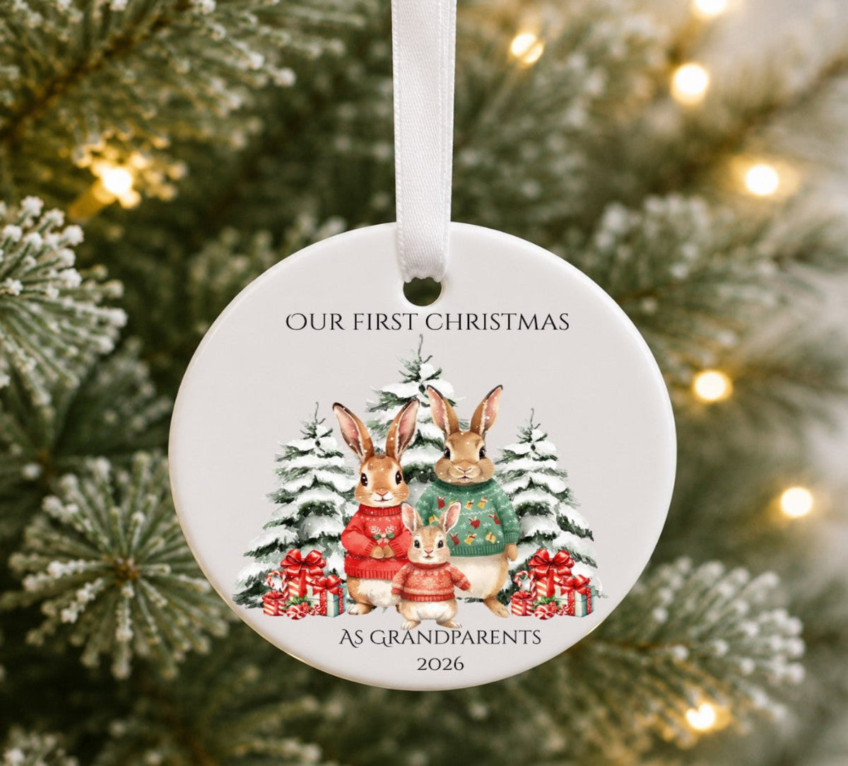 A round white Christmas ornament hanging from a white ribbon, featuring an illustration of three rabbits in winter sweaters with pine trees and gift boxes. The text reads 'OUR FIRST CHRISTMAS' at the top and 'AS GRANDPARENTS 2026' at the bottom.