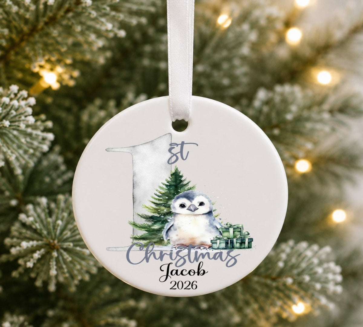 A personalised round ceramic Christmas ornament hanging from a white ribbon, featuring a watercolour penguin, evergreen tree, and gift box, with the text '1st Christmas Jacob 2026', set against a blurred Christmas tree background with warm lights.