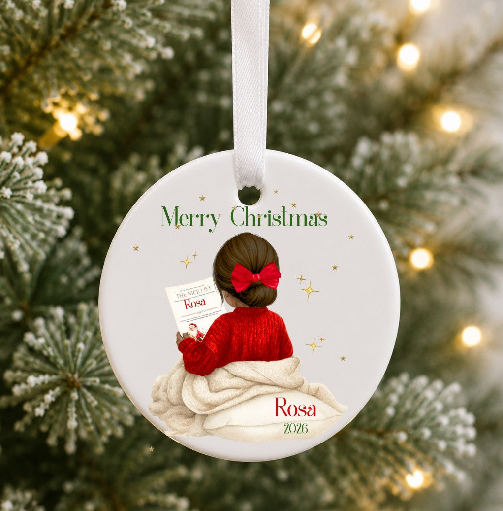 A personalised round white Christmas ornament hanging on a decorated Christmas tree. The ornament features an illustration of a woman with brown hair in a red bow and red sweater, wrapped in a beige blanket, reading a letter.