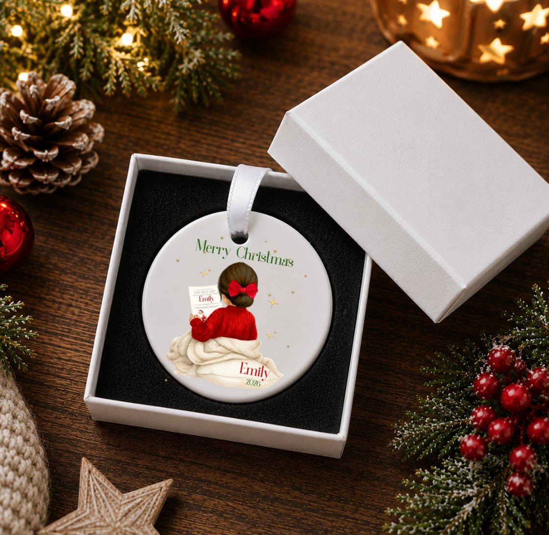 A round Christmas ornament displayed in an open white gift box on a dark wooden surface, surrounded by holiday decorations including pine branches, red berries, a pinecone, and warm string lights. The ornament features a cartoon illustration of a person in a red sweater and mask holding a gift, with the text 'Merry Christmas' and the name 'Emily' printed on it.