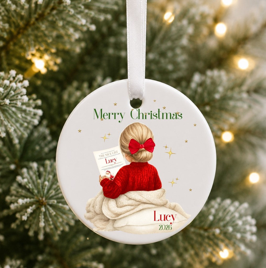A personalised round ceramic Christmas ornament hanging from a white ribbon, featuring an illustration of a blonde girl in a red sweater and red bow, wrapped in a blanket and holding a card that says 'Merry Christmas' and 'Lucy 2025'. The background shows a blurred Christmas tree with lights.