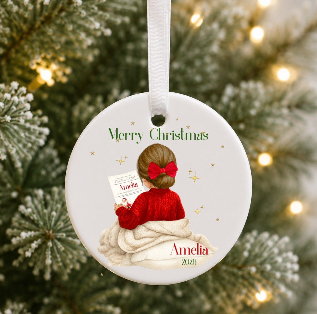 A personalised round ceramic Christmas ornament hanging from a white ribbon, featuring an illustration of a girl in a red sweater and red bow sitting on a blanket, with the text 'Merry Christmas' and 'Amelia 2025'. The ornament is photographed against a blurred background of a Christmas tree with warm lights.