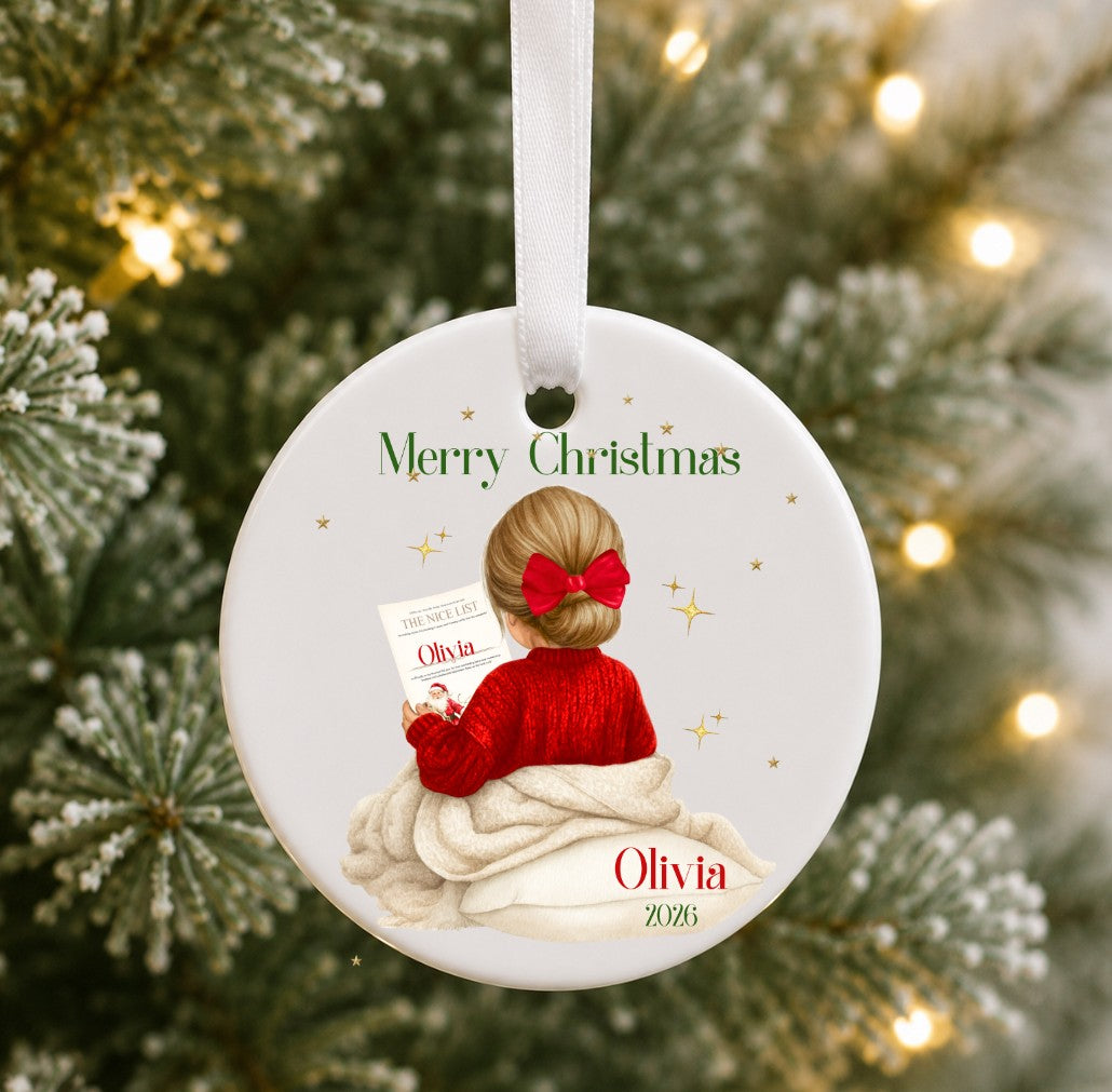 A personalised round ceramic Christmas ornament hanging on a Christmas tree. The ornament features an illustration of a girl with blonde hair in a red bow and red sweater, wrapped in a blanket and holding a card that says 'Olivia'.
